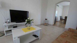 A living room with a flat-screen TV, a white coffee table with a vase of flowers, a sofa, and a dining area in the background.