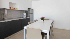 A modern kitchen with dark cabinets, a white countertop, and a small refrigerator. There is a dining table with chairs and a vase of flowers.