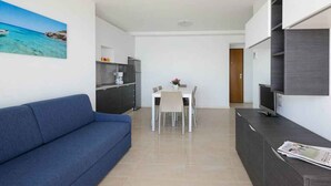 A modern living room with a blue sofa, a dining area with a white table and chairs, a kitchen with dark cabinets, and a TV on a wooden stand.
