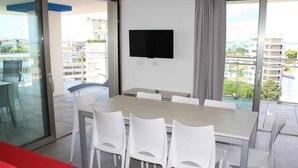 Un comedor moderno con una mesa grande y sillas blancas, un televisor de pantalla plana montado en la pared y un balcón con vistas a la ciudad.