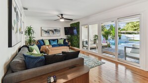 Una sala moderna con sofá, una mesa de centro y televisión. Hay un ventanal corredero que da a un área de piscina con mobiliario exterior.
