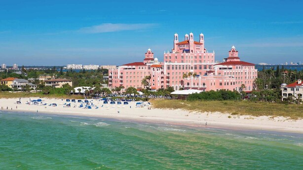 Un hotel grande y rosa con múltiples torres, una playa con sombrillas y un cielo azul despejado.