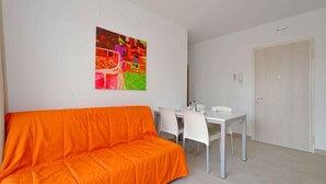 Une pièce avec un canapé orange vif, une table à manger blanche avec des chaises, et un tableau abstrait coloré accroché au mur.