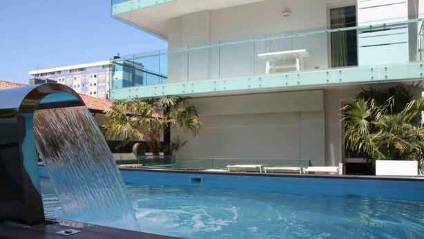 Une piscine agrémentée d’une cascade, de chaises longues et d’une balustrade en verre offrant une vue sur le paysage urbain.