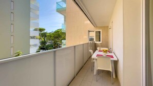 A balcony with a white table and chairs, a small window, and a view of buildings and trees. - Holiday apartment with pool and air conditioning (Lignano)