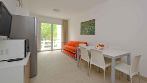 A modern living room with a sofa, a dining table with chairs, a TV, and a refrigerator. - Holiday apartment with pool and air conditioning (Lignano)
