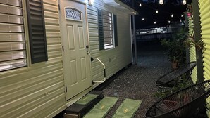 A small, single-story RV with a sliding door and a window with a decorative grille. There are steps leading to the door, and a chair is placed outside. - Tiny Wooden Home in Vega Baja with pool and close to the beach (Vega Baja)