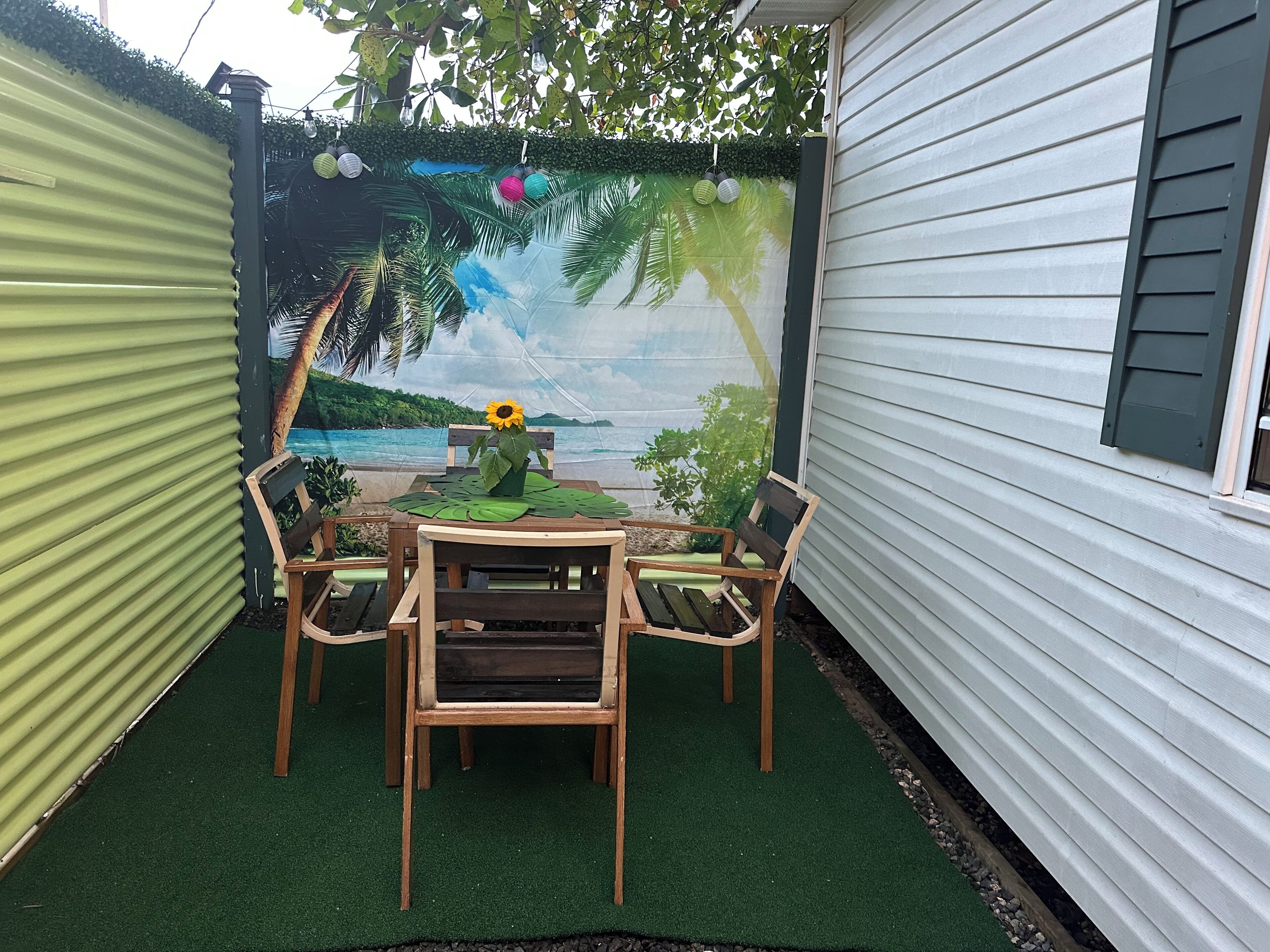 Eine Terrasse mit Tisch und Stühlen, ein Wandgemälde mit Strandmotiv und eine kleine Gartenanlage mit einer Sonnenblume.