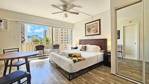 A hotel room with a bed, a desk, a chair, a ceiling fan, and a balcony with a view of buildings and palm trees. - Comfortable King Bed Waikiki Studio 2 mins walk to Beach with WiFi & AC (Honolulu)