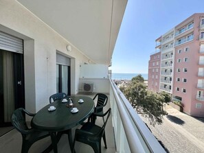 A balcony with a table and chairs, overlooking a coastal area with buildings and a clear sky.