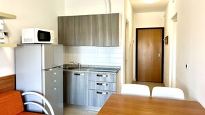 Una cocina compacta con microondas, refrigerador y un lavamanos pequeño. Hay una mesa de comedor con dos sillas y una puerta de madera al fondo.