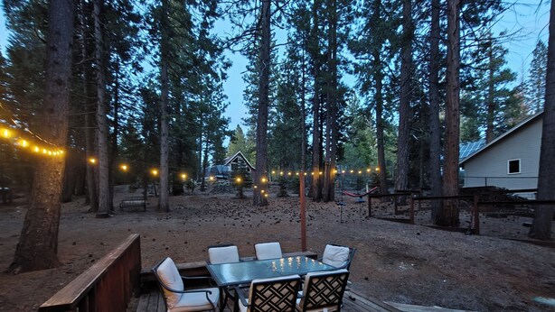 Une terrasse en bois avec une table et des chaises, entourée de grands arbres et avec une maison en arrière-plan.