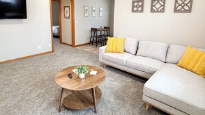 A living room with a sofa, a coffee table, and a TV. There are framed pictures on the wall and a door leading to another room. - 146 Wedgewood Wonder - Charming 2 bed 1 bath (Lincoln)