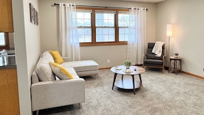 A living room with a sofa, a round coffee table, a chair, a lamp, and a window with curtains. - 146 Wedgewood Wonder - Charming 2 bed 1 bath (Lincoln)