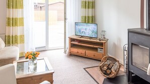 Un salon avec un téléviseur posé sur un meuble en bois, une cheminée, une table basse en verre ornée de fleurs et un panier en osier rempli de bois de chauffage.