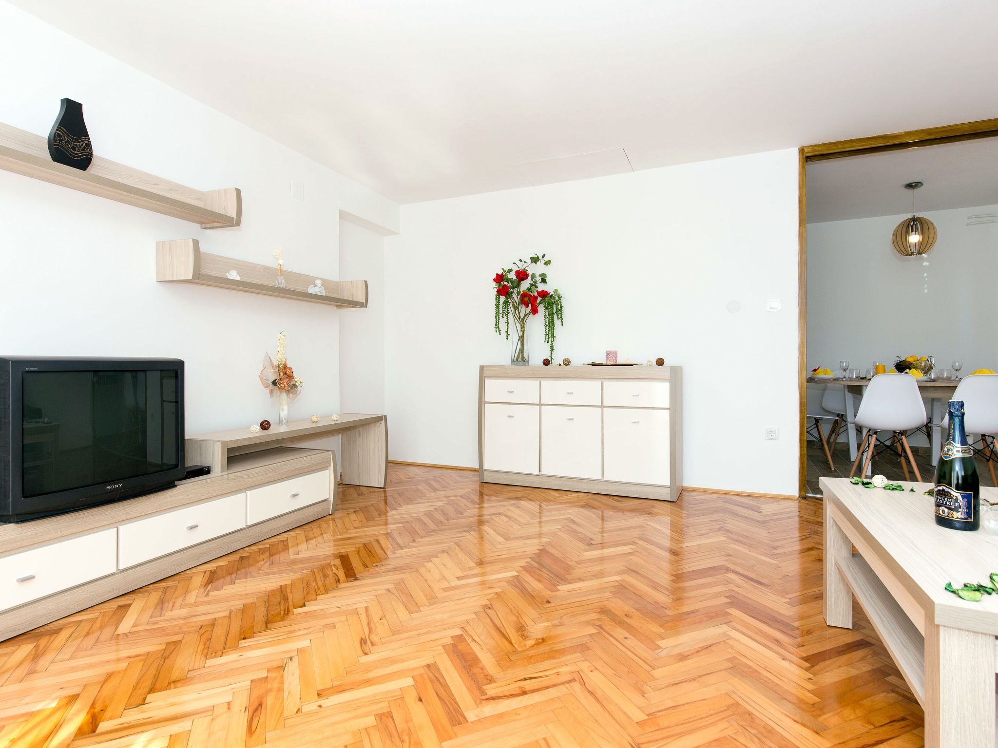 Un salon moderne avec un parquet en bois disposé en chevrons, un téléviseur à écran plat, un meuble blanc et un coin repas visible à travers une porte ouverte.