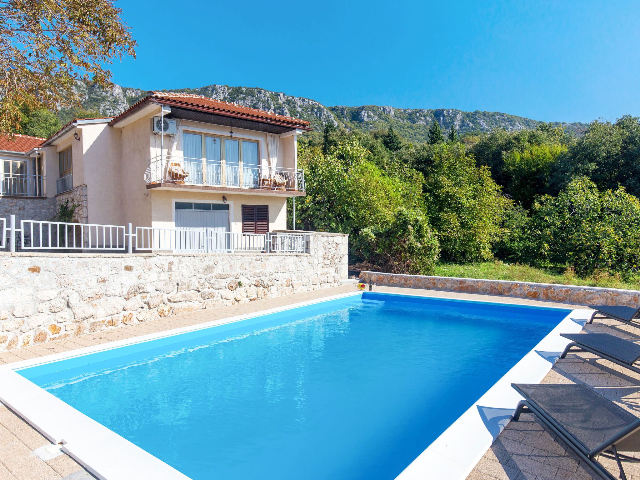 A poolside view of a two-story building with a stone wall, a balcony with outdoor furniture, and a mountainous backdrop.