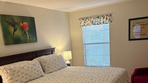 A neatly made bed with a floral-patterned bedspread, a nightstand with a lamp, a window with blinds, and a framed sign on the wall. - "Nothing But Sunshine" (Fort Myers)
