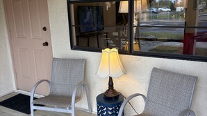 A seating area with two chairs and a table with a lamp, a door, and a window with blinds. - "Nothing But Sunshine" (Fort Myers)