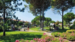 Um parque com grama verde, árvores e pessoas. Há bancos e uma fonte.