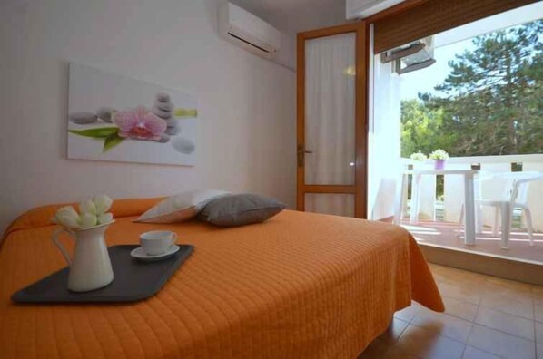 A bedroom with an orange bedspread, a painting of flowers on the wall, a white pitcher and cup on a tray, and a view of an outdoor patio with chairs.