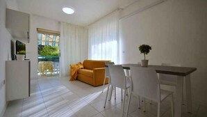 Un salon moderne et bien éclairé, avec un canapé, une table à manger et des chaises. On peut voir une plante en pot sur la table, ainsi qu’une fenêtre avec des rideaux.