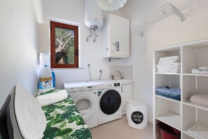 A laundry room with a washing machine, dryer, ironing board, and a shelf with folded towels.