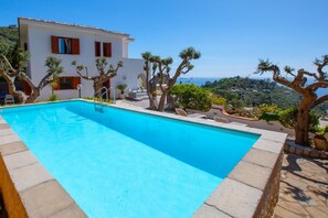 A swimming pool with a clear view of the sea, a white building with red shutters, and a landscaped garden with trees.