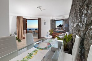 A modern hotel room with a dining area, a view of the sea, a stone wall, and a ceiling fan.