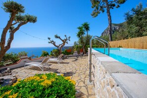 A stone pool with a clear view of the sea, surrounded by lounge chairs and greenery.
