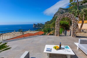 Una terraza con un arco de piedra, una mesa con una revista, una botella y limones, con vistas a un paisaje costero y cielo despejado.