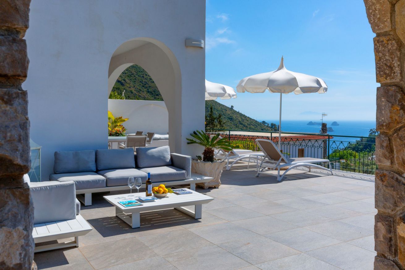 Une terrasse avec un canapé, des chaises, une table sur laquelle se trouve un saladier rempli de fruits et des parasols.