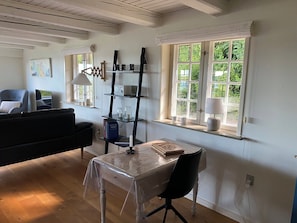Une pièce avec un plancher en bois, une table recouverte d’une vitre transparente, une chaise noire, une bibliothèque et une fenêtre donnant sur un paysage verdoyant.