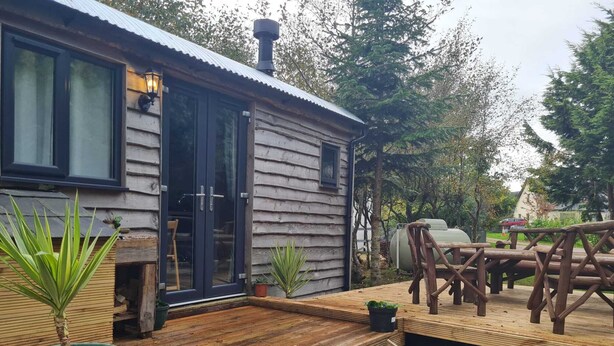 Une cabane rustique dotée d’une terrasse en bois, d’une porte noire et d’un petit espace extérieur aménagé pour s’asseoir.