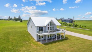 Une maison à deux étages avec un balcon, entourée d’un champ verdoyant et sous un ciel dégagé.