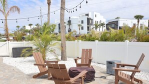 Un espace extérieur aménagé avec des chaises en bois et un banc couvert, entouré de palmiers et d’une clôture blanche.