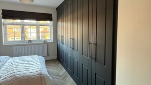 Une chambre dotée d’une grande armoire, d’un lit avec une couette blanche, d’un radiateur et d’une fenêtre donnant sur des immeubles.