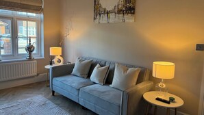 Un salon chaleureux avec un canapé gris, deux tables d’appoint avec des lampes, un tableau au mur et une vue par la fenêtre.