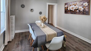 Een eetkamer met een lange tafel, witte stoelen, een vaas met gedroogde bloemen en een bloemistisch schilderij aan de muur.