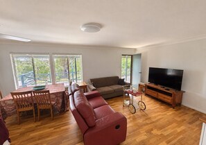A living room with a sofa, a TV, a dining table with chairs, and a window with a view of greenery.