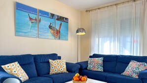 Een woonkamer met twee blauwe banken, gestreepte kussens, een witte salontafel met een fruitschaal, en een schilderij van boten aan de muur.