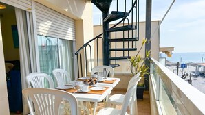 Een balkon met een tafel gedekt voor twee personen, witte plastic stoelen, een spiraaltrap en uitzicht op het strand.