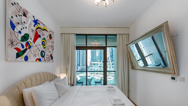 A hotel room with a large bed, a view of a cityscape through a window, and a colorful abstract painting on the wall. - 1BR Apt in Prime Location w/ Marina View Balcony (Dubai)