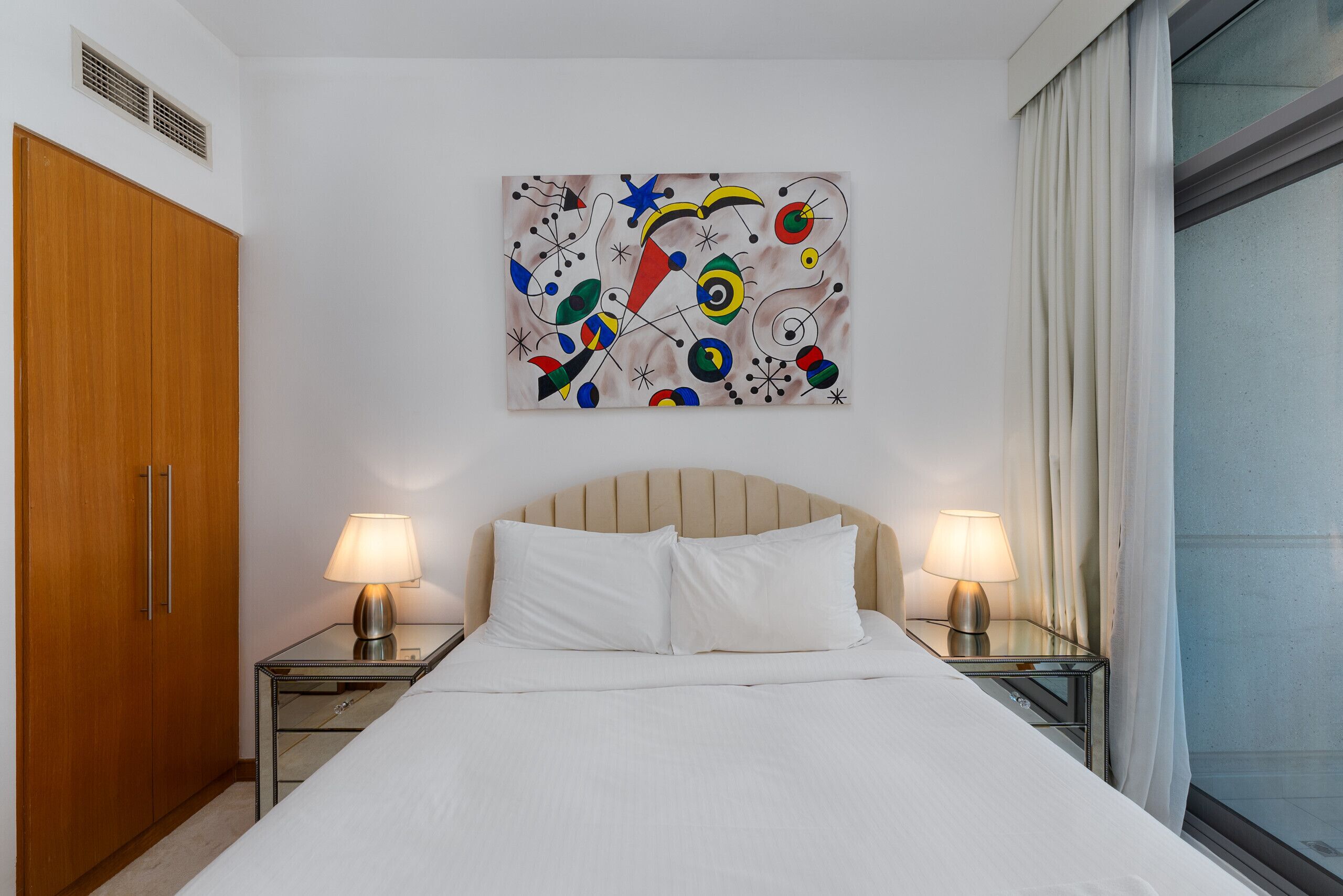 Une chambre d’hôtel avec un lit, deux tables de chevet avec des lampes, une armoire en bois et un tableau abstrait aux couleurs vives accroché au mur.