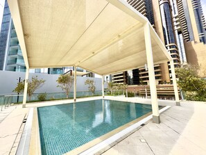 A swimming pool with a tiled deck, surrounded by modern buildings and a covered seating area. - 1BR Apt in Prime Location w/ Marina View Balcony (Dubai)