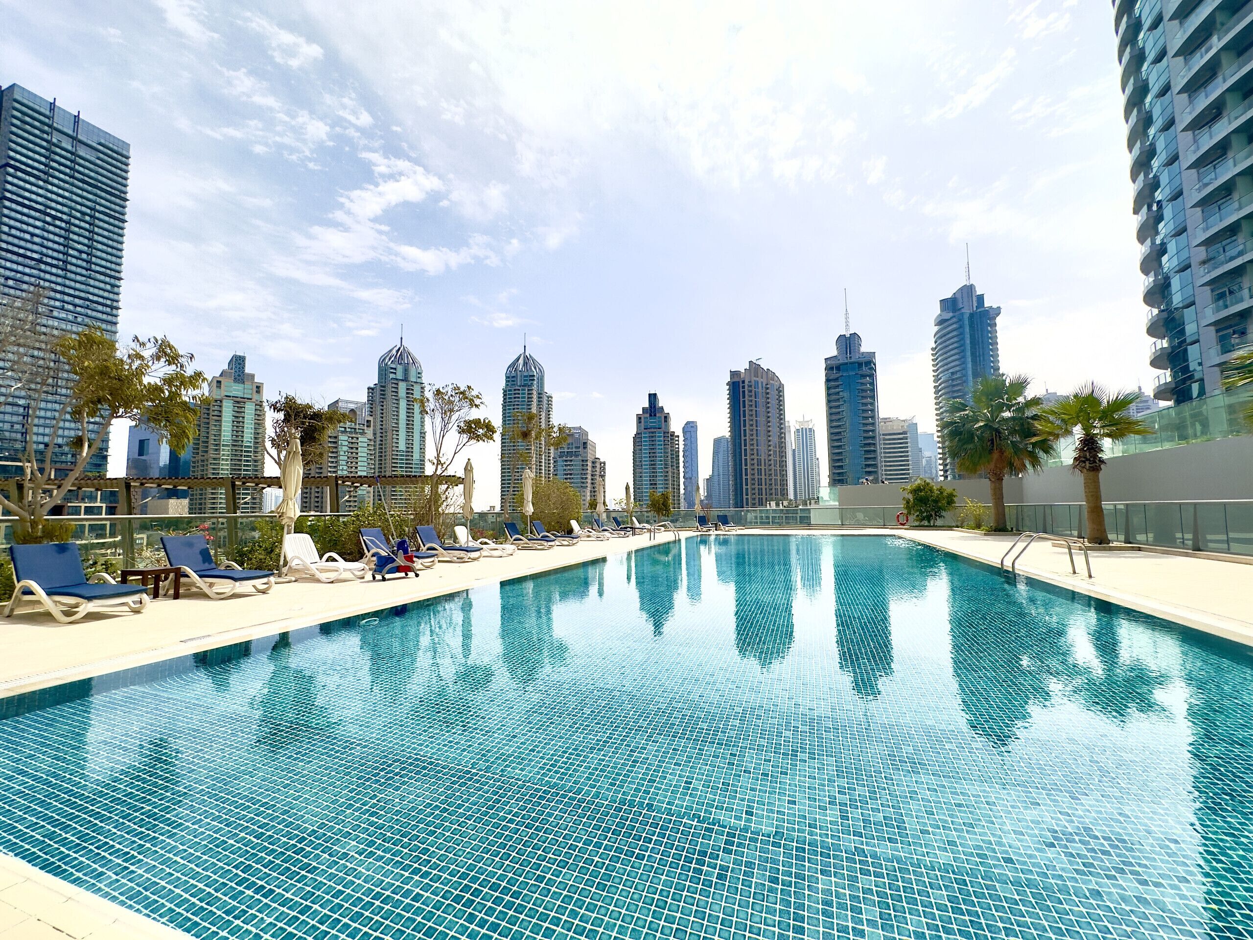 A rooftop pool with lounge chairs and palm trees, surrounded by high-rise buildings.