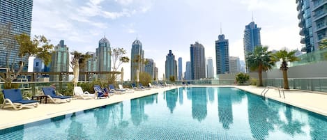 Una piscina en la azotea con sillones de descanso y palmeras, rodeada de edificios altos.