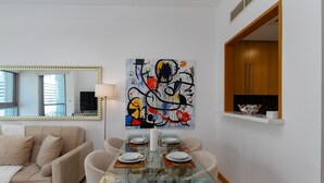 A modern living room with a glass dining table set for four, a beige sofa, a large abstract painting, and a wooden cabinet. - 1BR Apt in Prime Location w/ Marina View Balcony (Dubai)