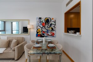 A modern living room with a glass dining table set for four, a beige sofa, a large abstract painting, and a wooden cabinet. - 1BR Apt in Prime Location w/ Marina View Balcony (Dubai)