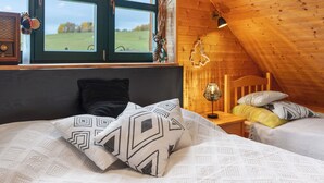 Een knus slaapkamer met een houten plafond, een groot raam, een bed met gestreepte kussens, en een nachtkastje met een lamp.